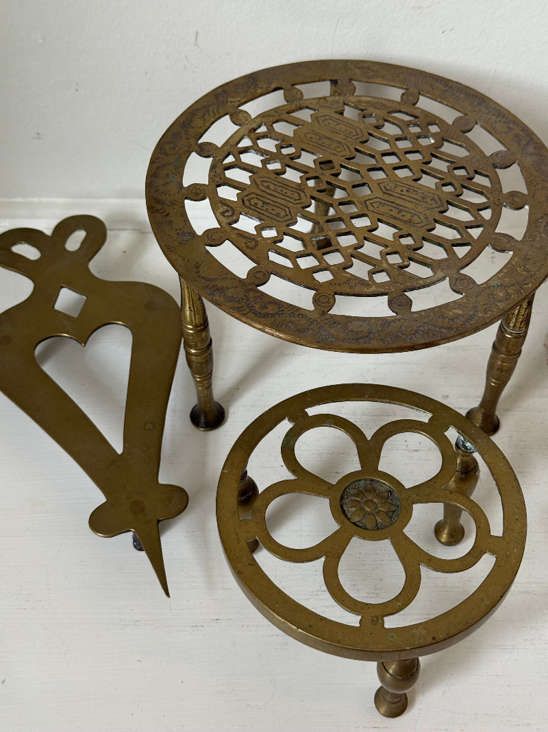 Vintage brass trivet trio with ornate cutwork designs and aged patina, photographed on a white surface