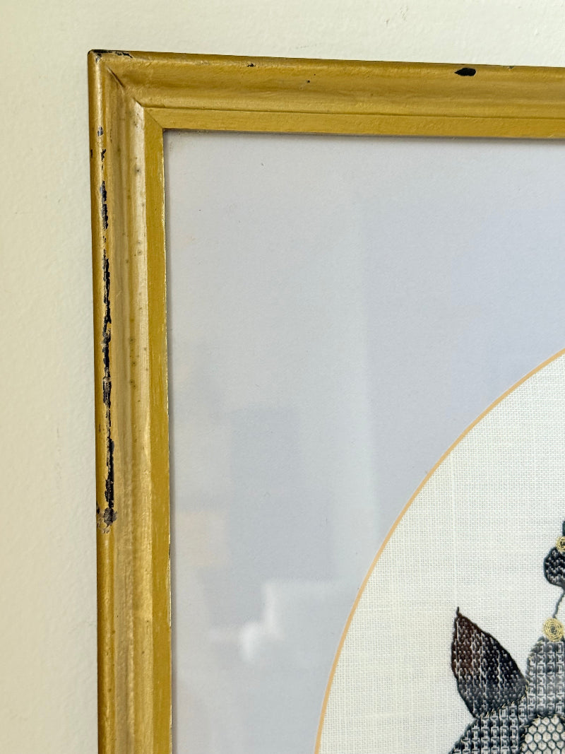 Framed circular hand embroidery of birds and fuchsia flowers in black and grey on white linen, set in gold frame, white surface background.
