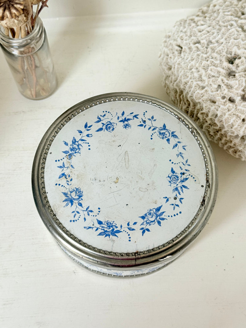Vintage round shortbread tin with blue floral pattern on silver background, photographed on a white surface.