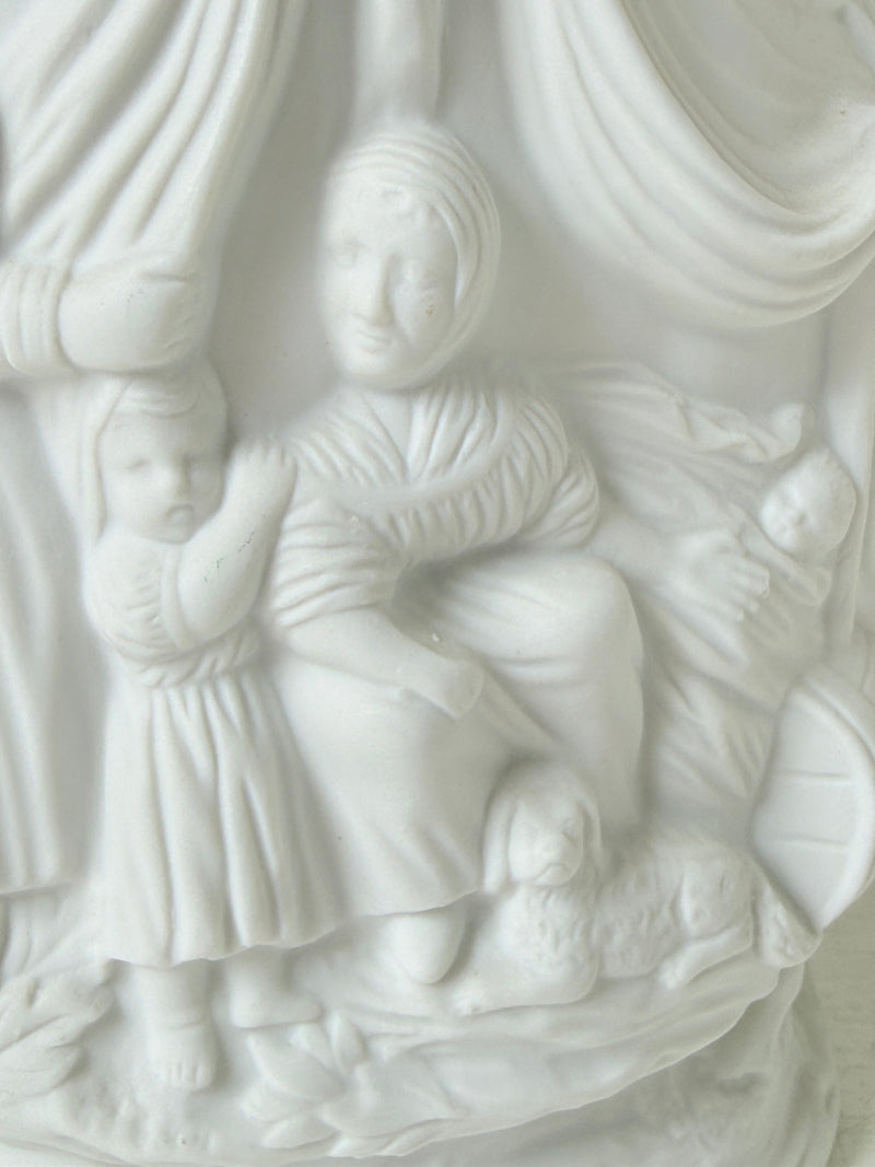 A close-up of a white sculptural relief featuring a seated woman, a standing child, and a baby, with draped fabric and soft carved details.