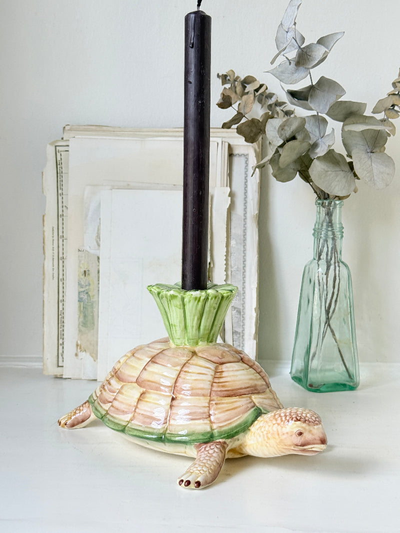 A ceramic turtle candle holder with a green fluted base holding a black taper candle, styled on a white surface with a green glass vase of eucalyptus and framed art in the background.