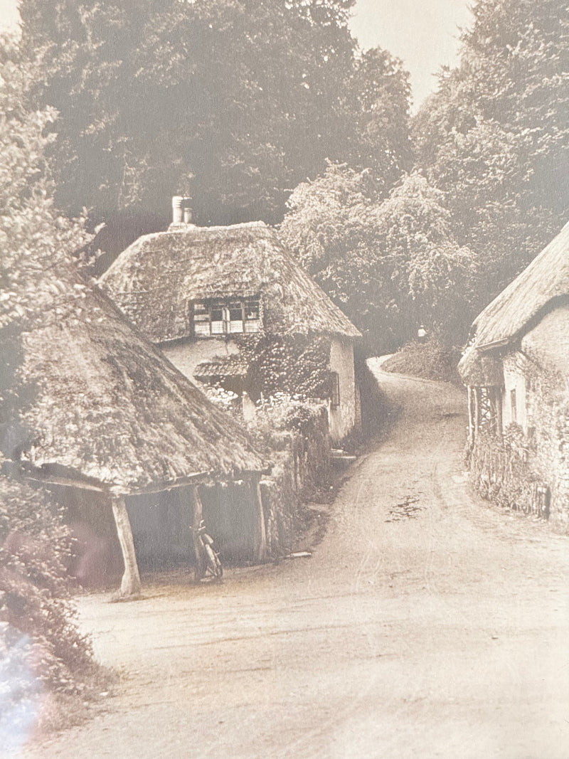 Framed antique sepia photograph of English village with cottages and trees, photographed on white surface with vase