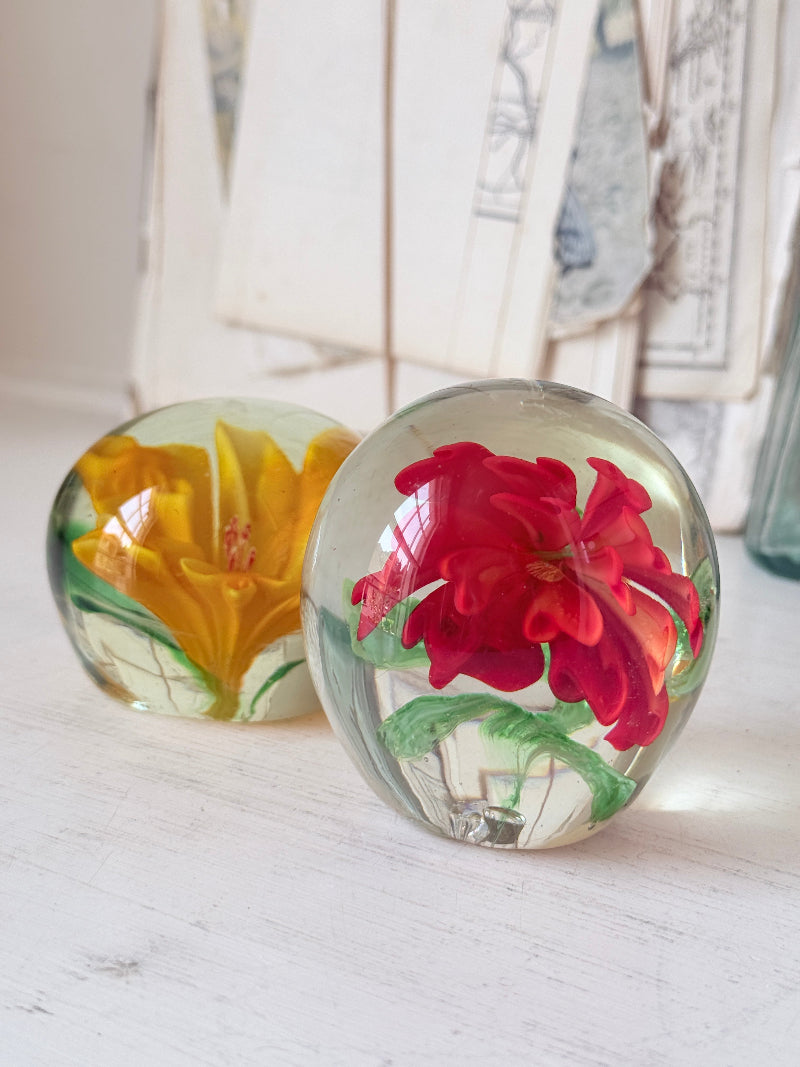 Pair of orange and yellow flower glass paperweights with green leaves, photographed on a white surface