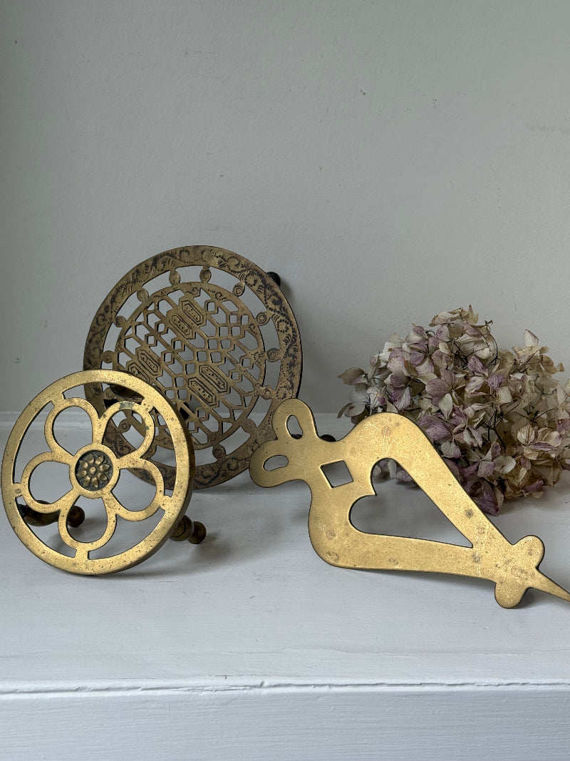 Vintage brass trivet trio with ornate cutwork designs and aged patina, photographed on a white surface