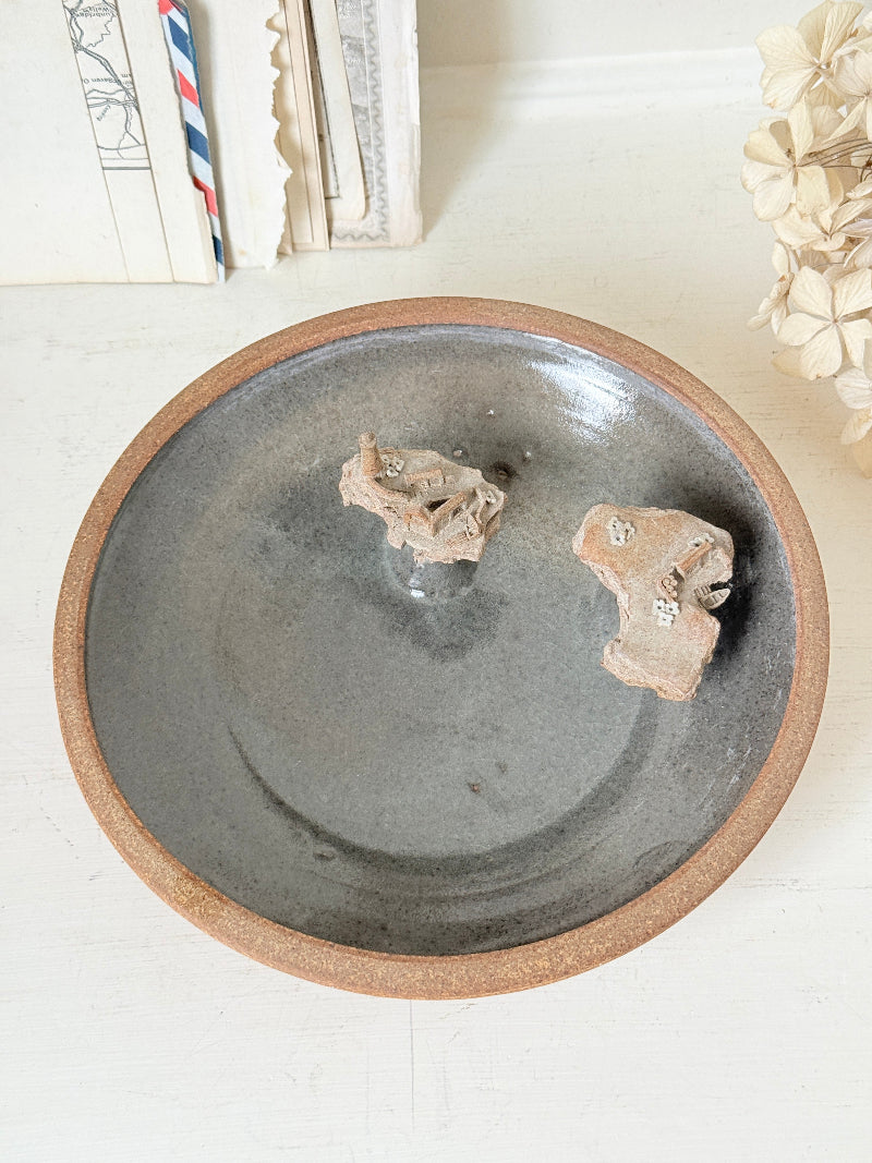A round ceramic shallow dish with a matte dark gray interior and a textured terracotta brown rim, holding two small beige decorative objects, set on a light surface with soft background decor.