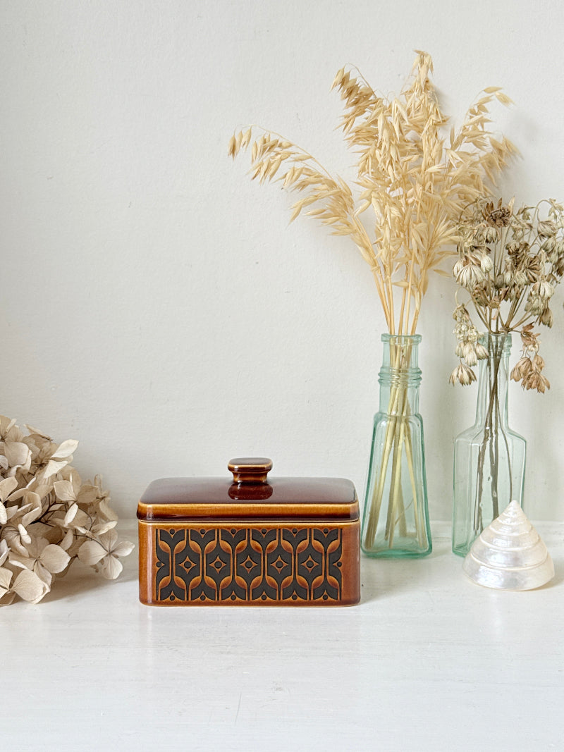 A rectangular ceramic lidded box with a glossy reddish-brown top and a dark brown geometric patterned front, styled on a white surface with dried flowers and glass vases against a white wall.