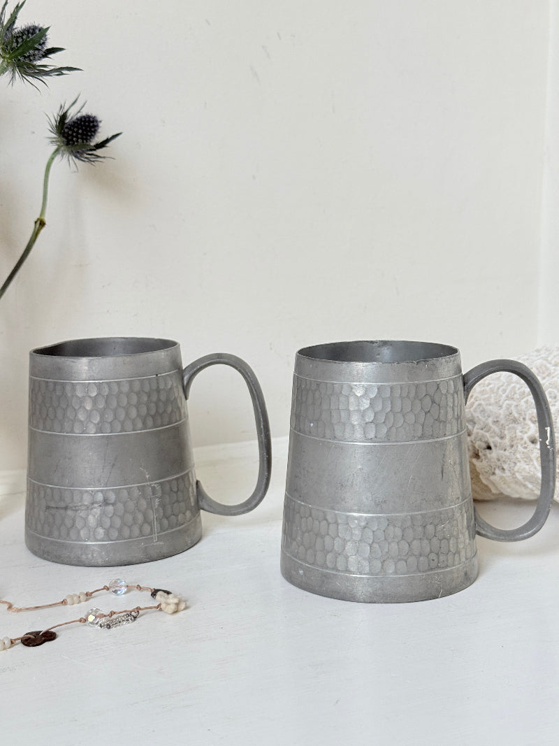 Pair of vintage pewter tankards with handles and textured detail, photographed on a white surface