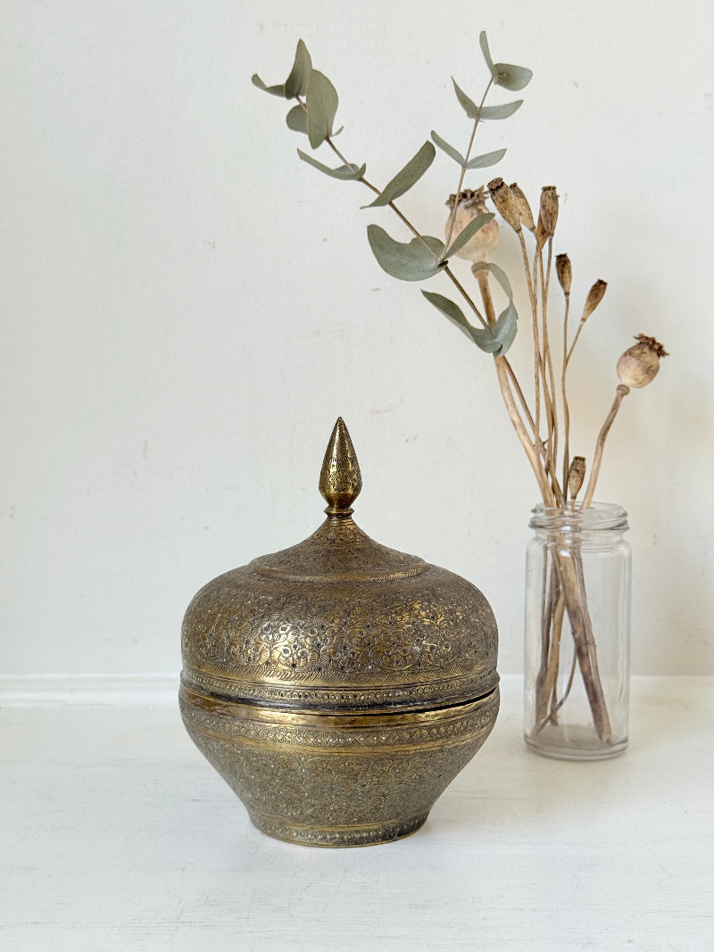 Indian Benares brass lidded jar with pointed finial and darkened interior, photographed on a white surface
