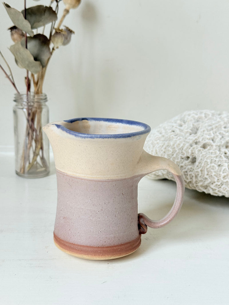 Studio pottery milk jug in cream and lavender glaze with impressed initials, photographed on a white surface.