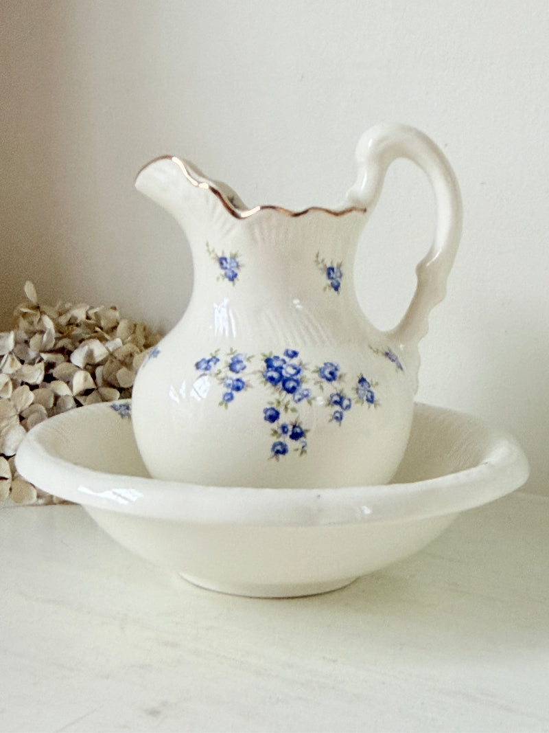 A white ceramic pitcher with blue floral patterns and a gold rim, displayed on a matching white ceramic bowl against a neutral cream background.