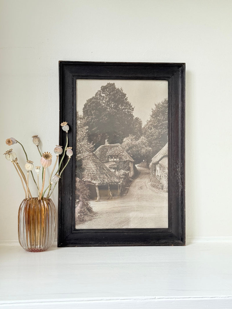 Framed antique sepia photograph of English village with cottages and trees, photographed on white surface with vase