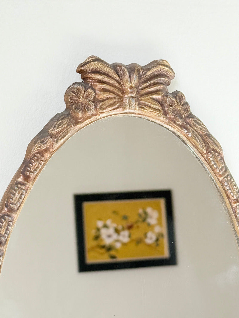 Vintage oval wall mirror with decorative gilt frame and bow crest, styled against a neutral wall with hydrangeas.