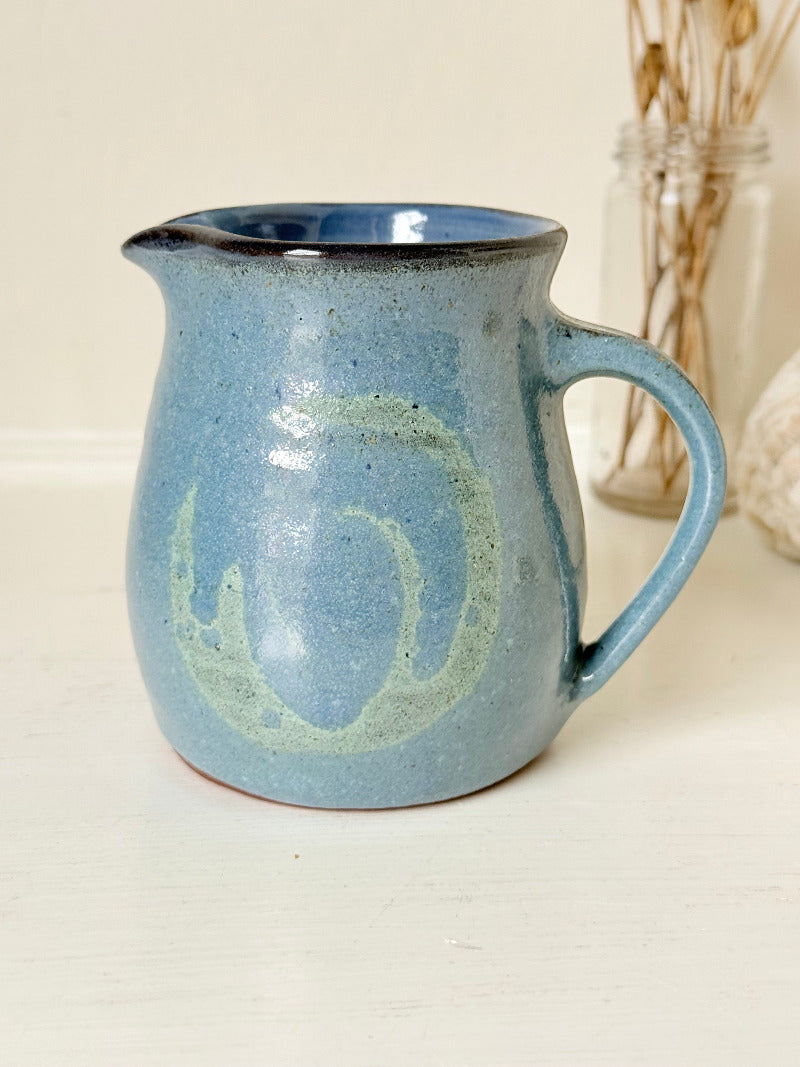 Vintage studio pottery milk jug with blue and green glaze, handmade with a rounded form and sturdy handle, styled on a white surface.