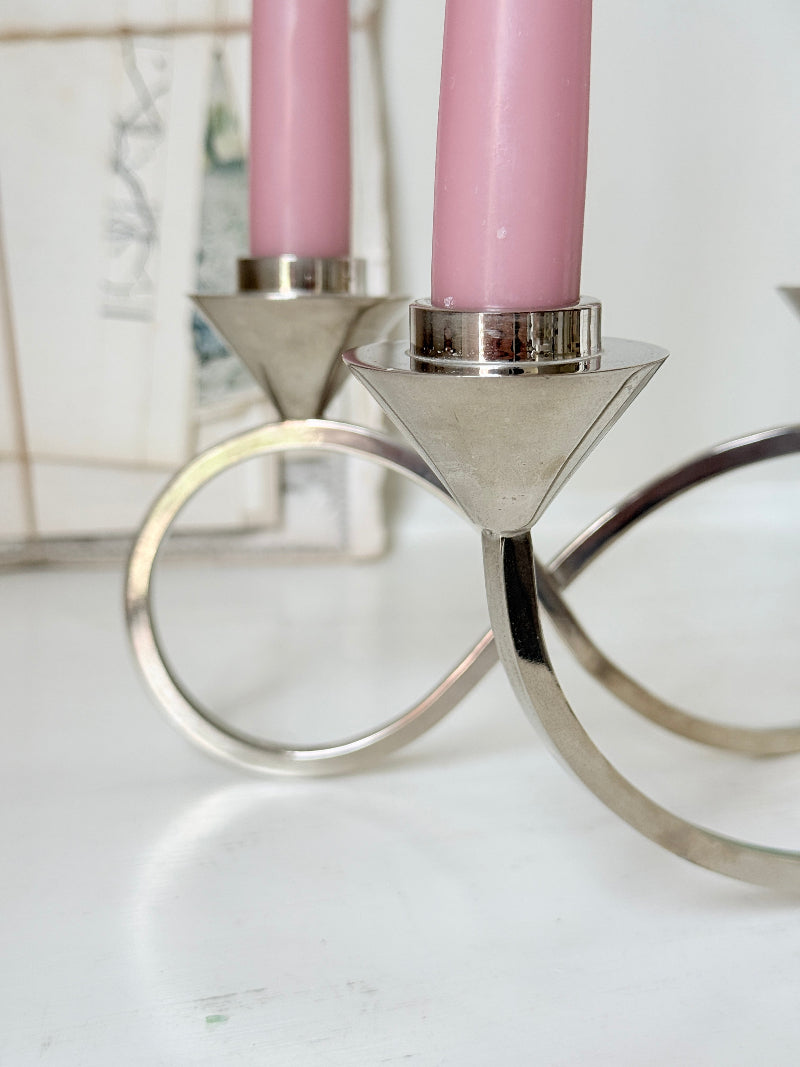 Silver tone five light candelabra with a sculptural looped base holding pink candles, photographed on a white surface