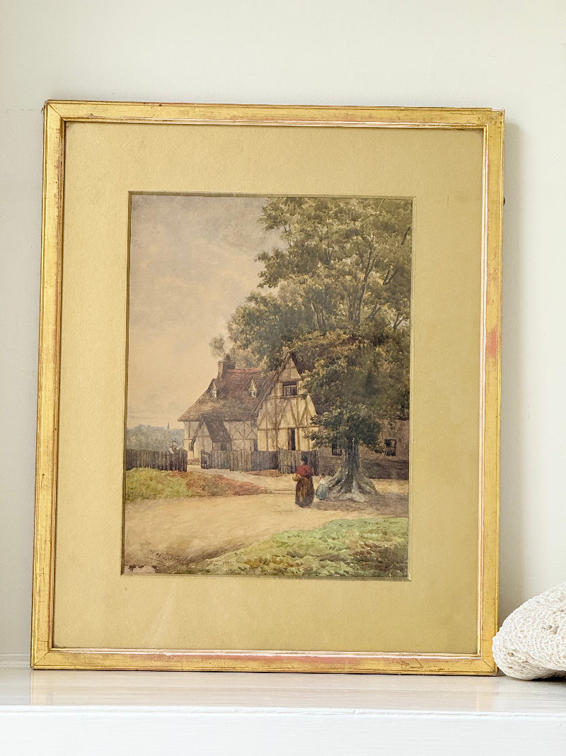 Antique framed watercolour of English village street with thatched cottages and figures, in gilt frame with wide mount, on white surface.
