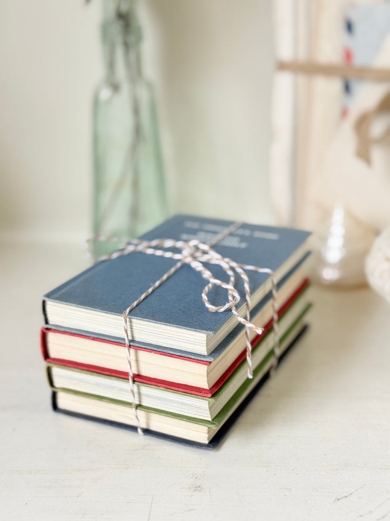 A stack of four small hardcover notebooks tied with white burlap twine, set on a light wooden surface with a softly blurred background.