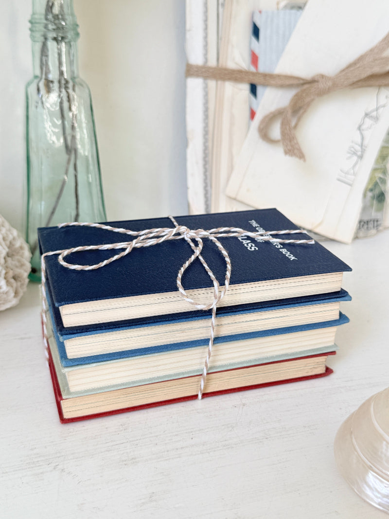 A stack of four hardcover books tied with white string, featuring navy, blue, light blue, and red covers, styled on a white wooden surface with a glass bottle and vintage papers in the background.