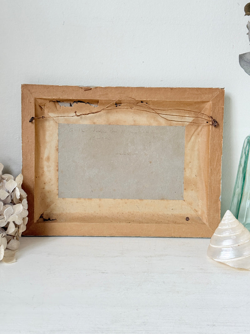 A rustic, vintage-style rectangular wooden picture frame shown from the back, resting on a white surface against a white wall, flanked by decorative seashells and a glass vase.