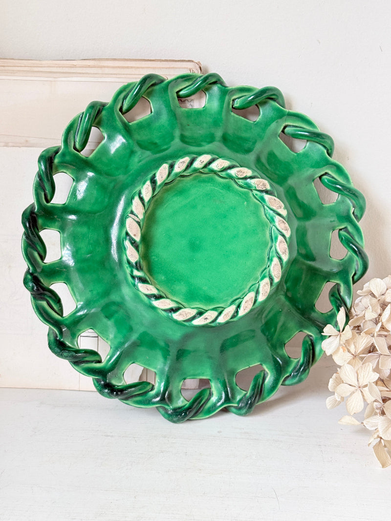 A green ceramic decorative plate with a scalloped, cutout rim and a twisted rope detail around the inner edge, displayed against a light background with dried flowers at the side.