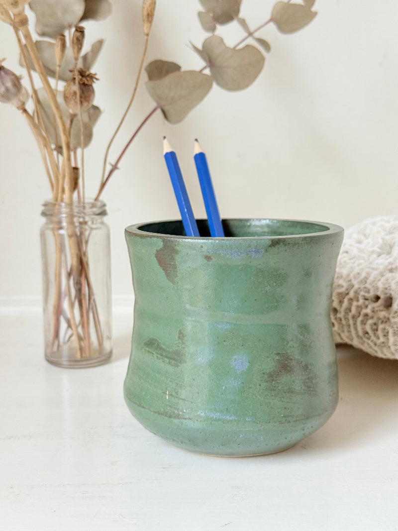 Studio pottery pencil pot in green glaze, handmade on the wheel, photographed on a white surface with pencils inside.