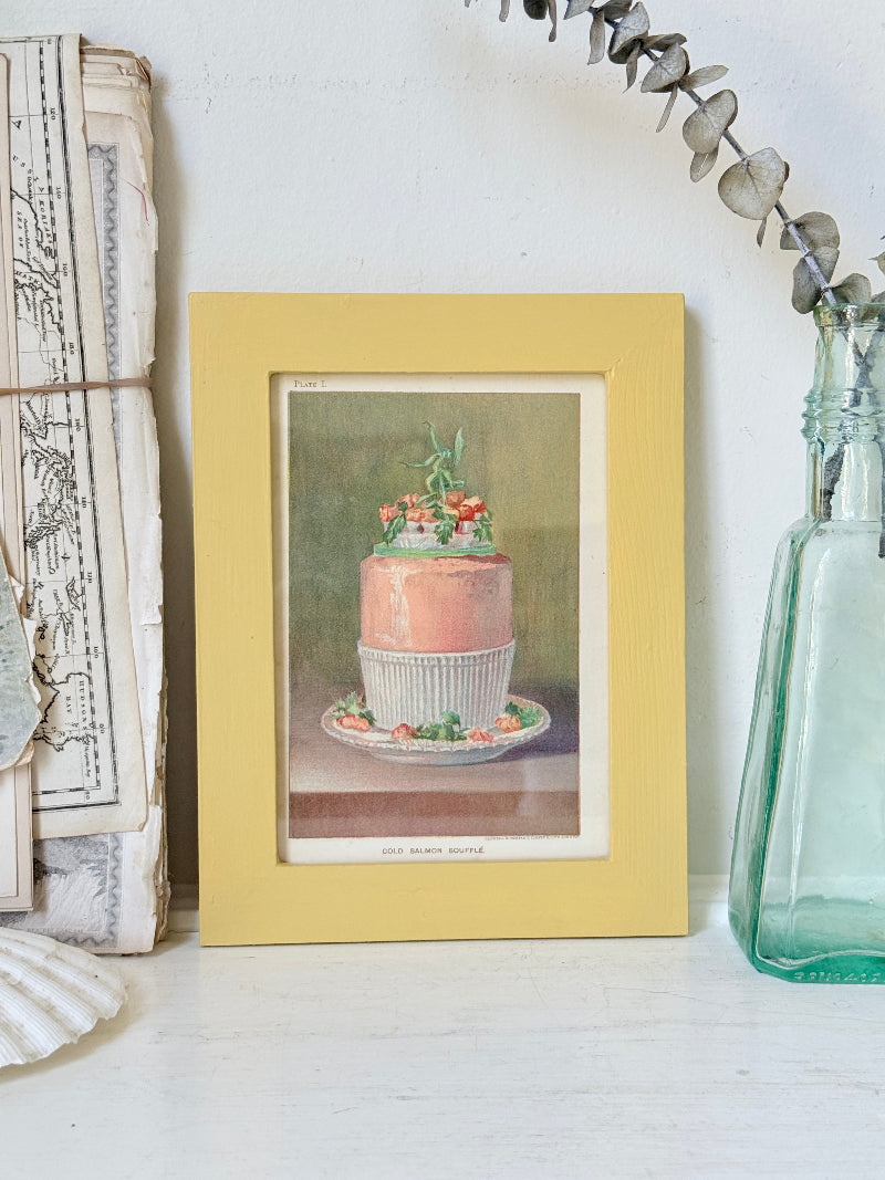 Framed Victorian cookery illustration of a celebration cake from 1896 in a mustard yellow frame displayed on a white surface.