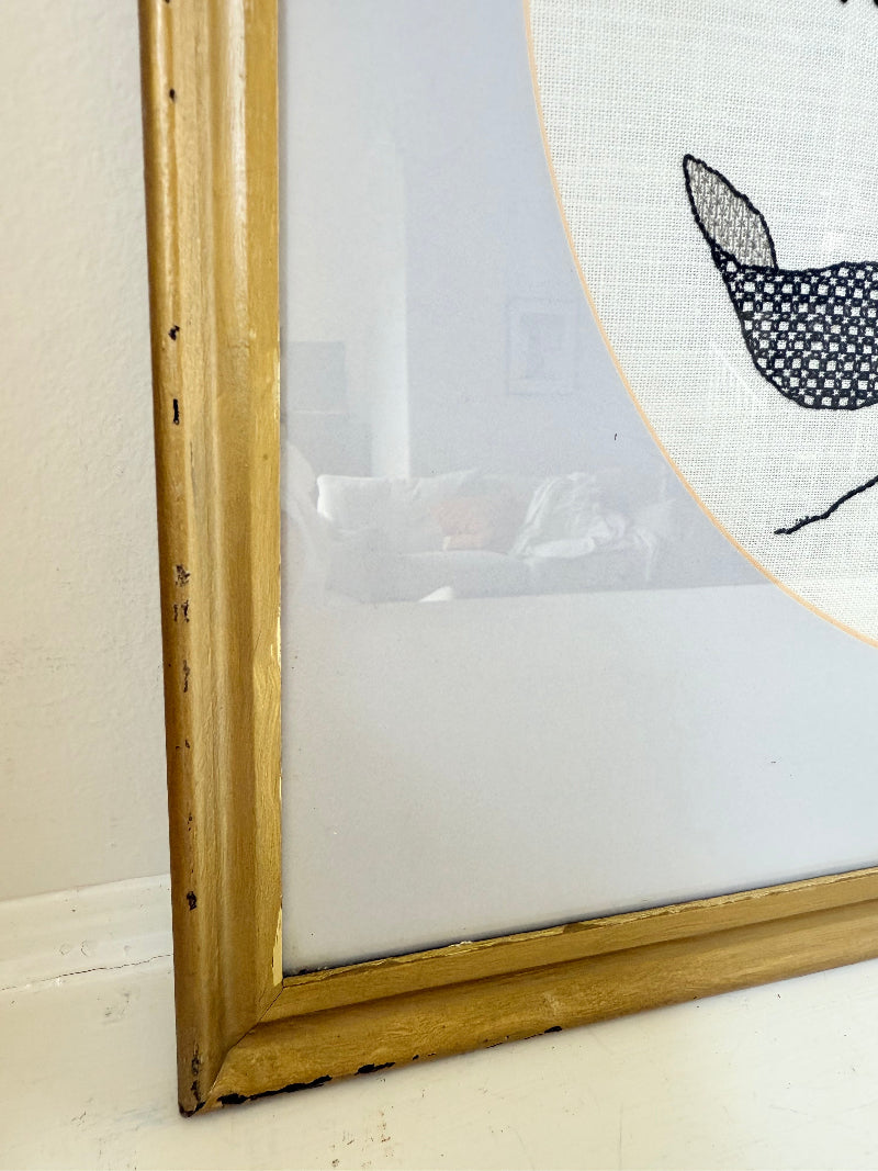 Framed circular hand embroidery of birds and fuchsia flowers in black and grey on white linen, set in gold frame, white surface background.