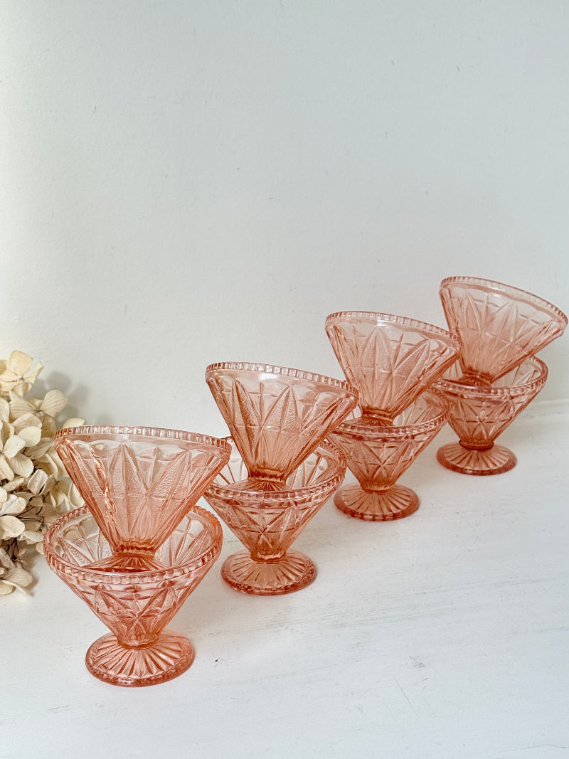 Six pink cut-glass cocktail glasses arranged on a white surface against a white background, with a small dried floral arrangement at the left edge.