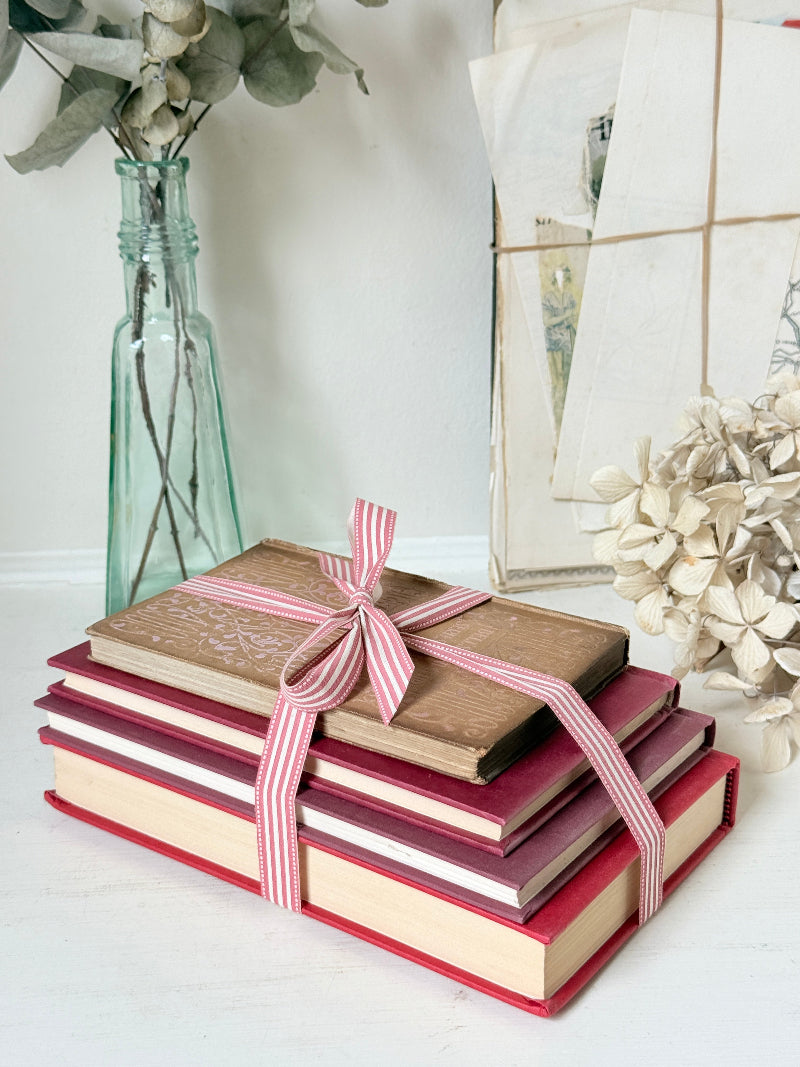 Stack of cloth bound vintage books in red and warm pink tones tied with striped ribbon and dried flowers