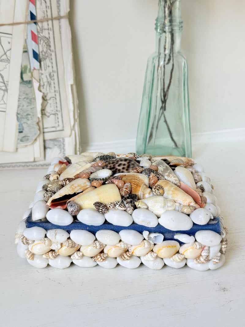 A rectangular decorative box or tray covered with white pebbles and assorted seashells, placed on a white tabletop with a tall translucent glass bottle behind it.