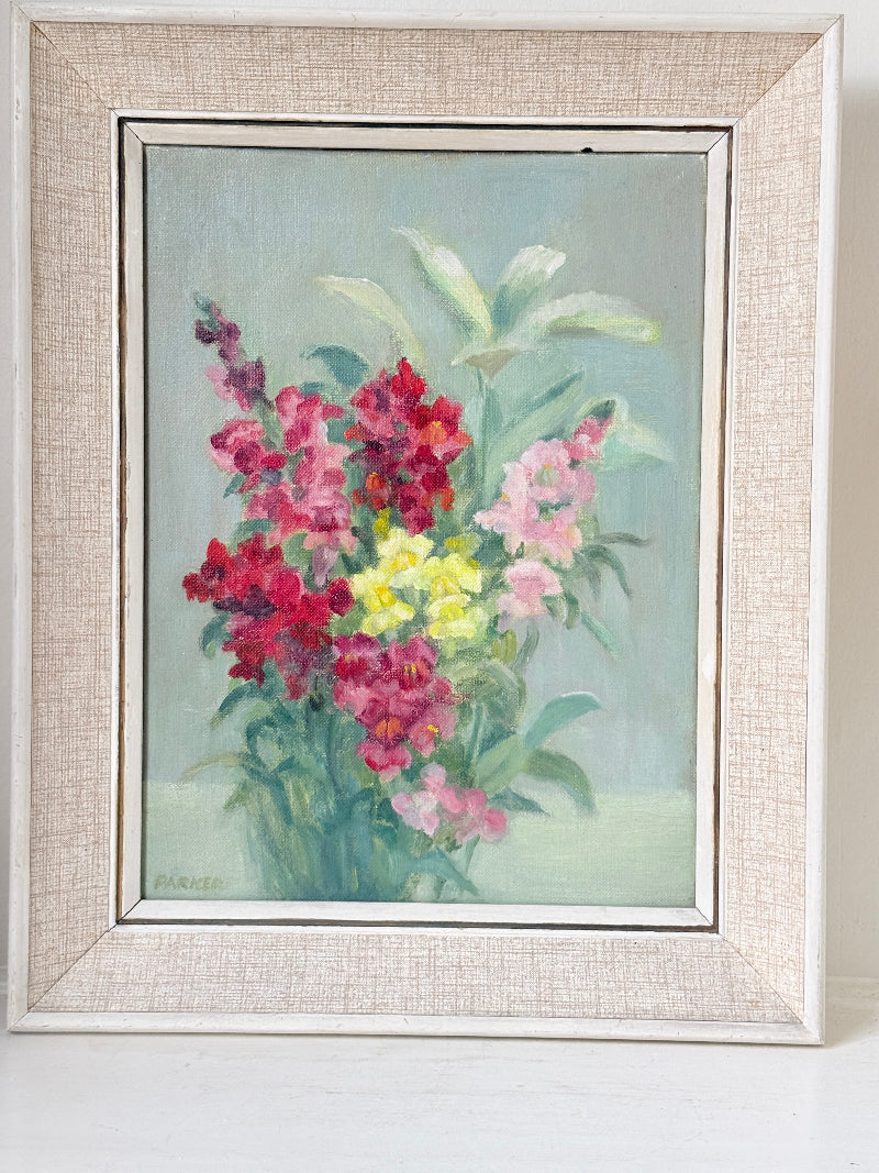 A framed floral painting featuring pink, red, and yellow flowers with green leaves on a soft blue-green background, housed in a distressed off-white textured frame.