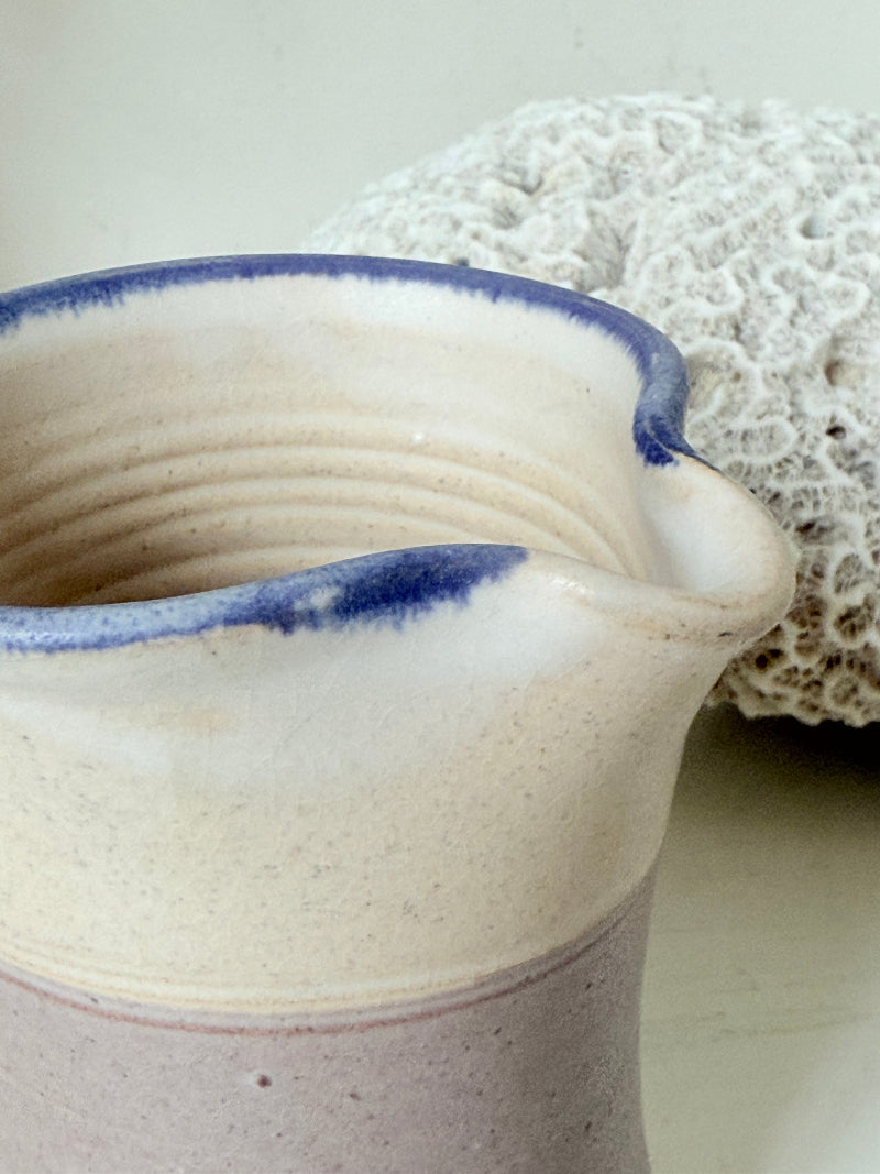 Studio pottery milk jug in cream and lavender glaze with impressed initials, photographed on a white surface.