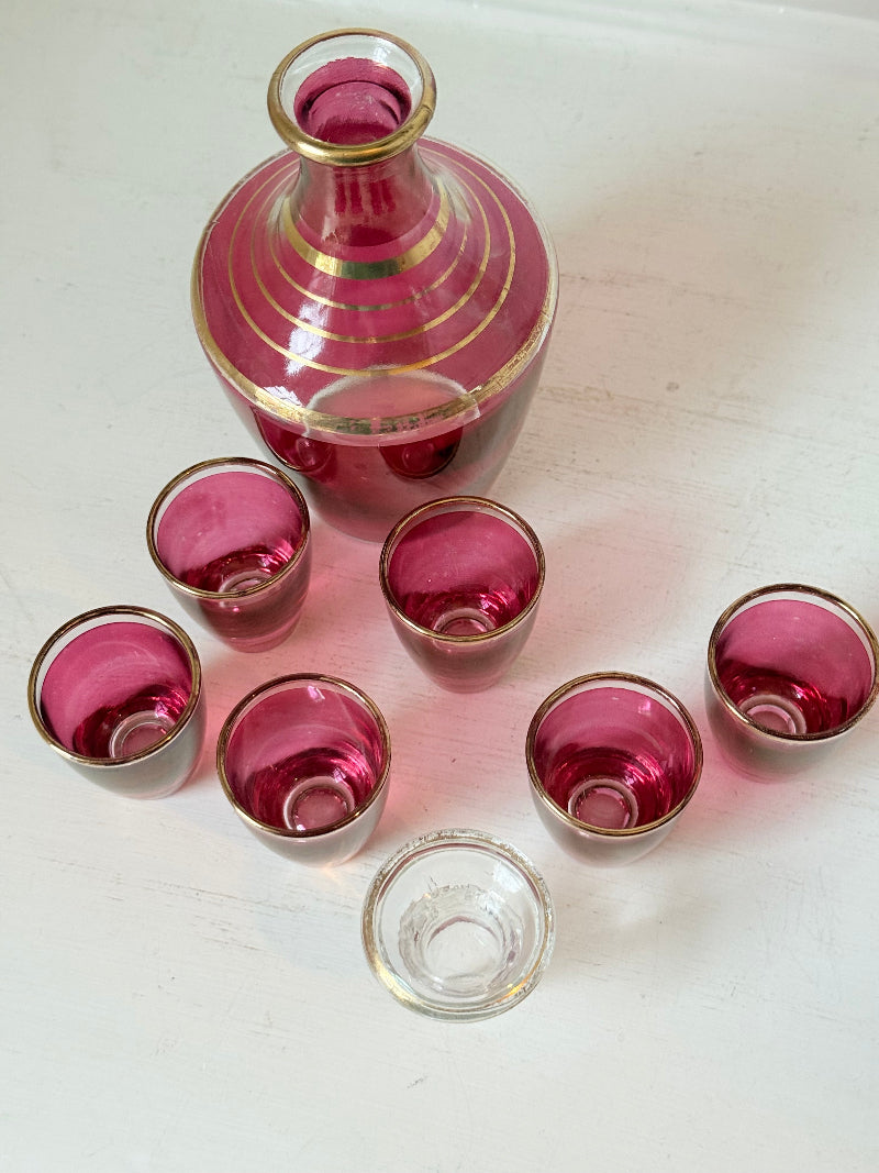 Vintage red printed glass cocktail set with clear glass decanter and six small glasses with gilt band rims, photographed on a white surface