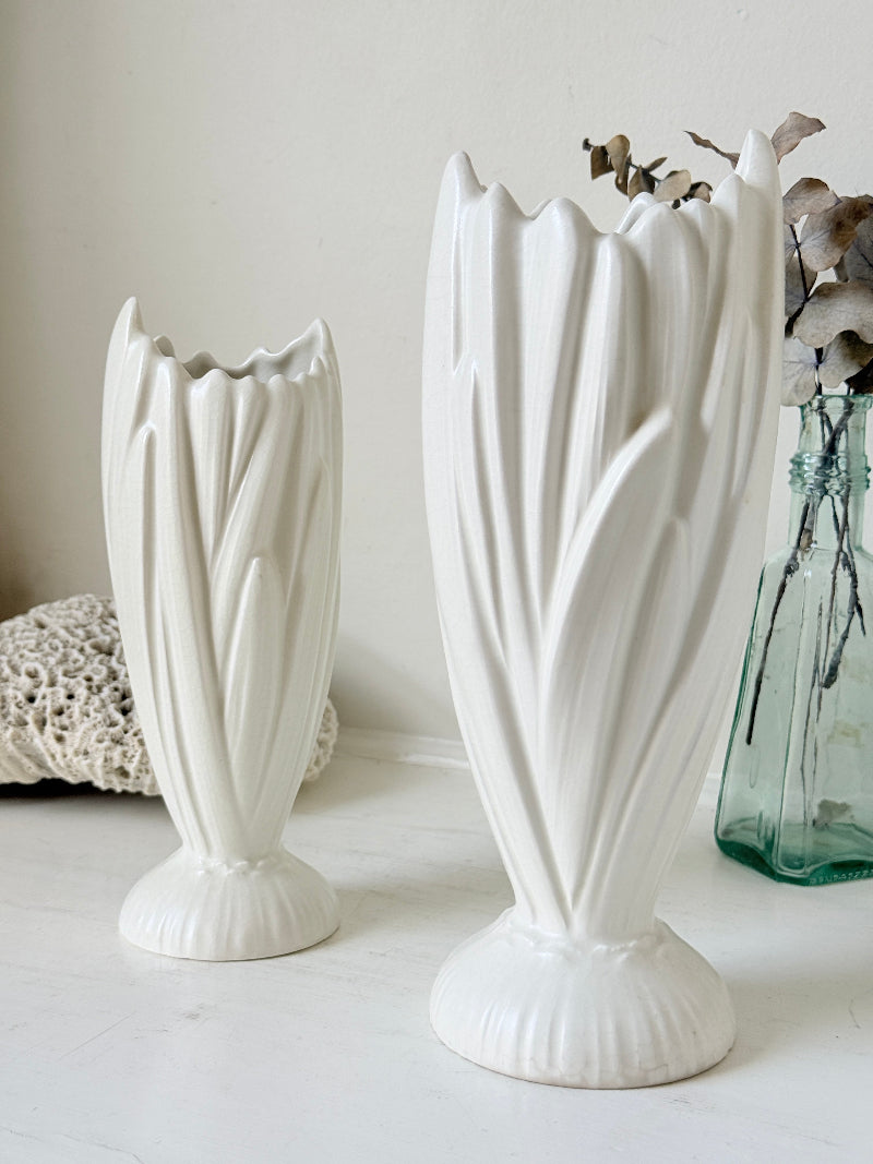 Two white ceramic tulip-shaped vases with fluted stems and scalloped rims, displayed on a white surface with a green glass vase and dried flowers in the background.