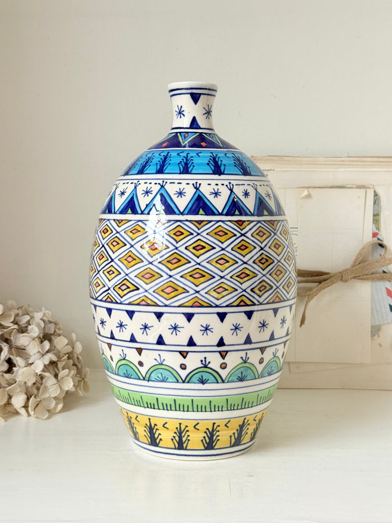 A ceramic vase with a wide bulbous body and narrow neck, decorated with multiple colorful geometric bands including blue, yellow, green, and white. It is photographed on a white surface against a light neutral background with dried flowers and a box as styled props.