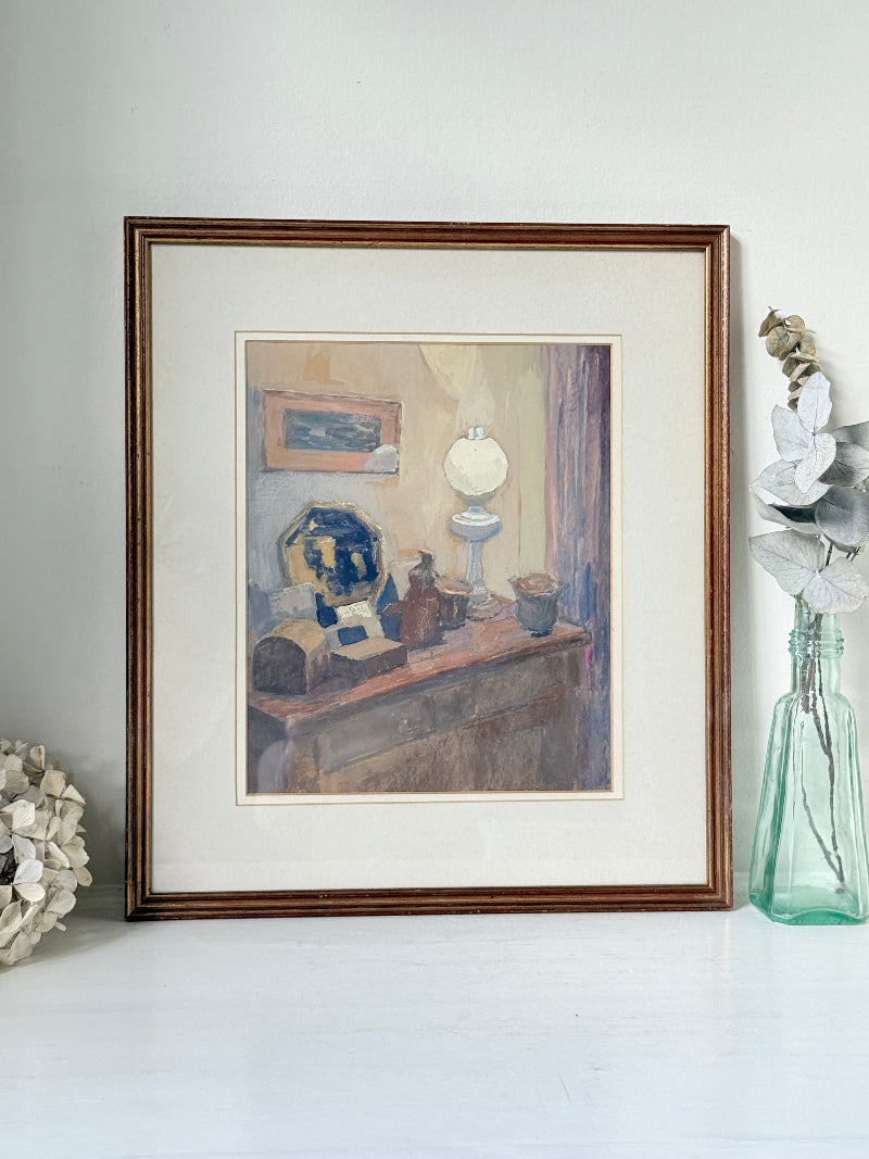 A framed still life painting displayed on a white surface against a light gray wall. The artwork depicts a tabletop scene with a blue-and-white vase, a white globe lamp, and small bottles, enclosed in a white mat and a brown wooden frame.