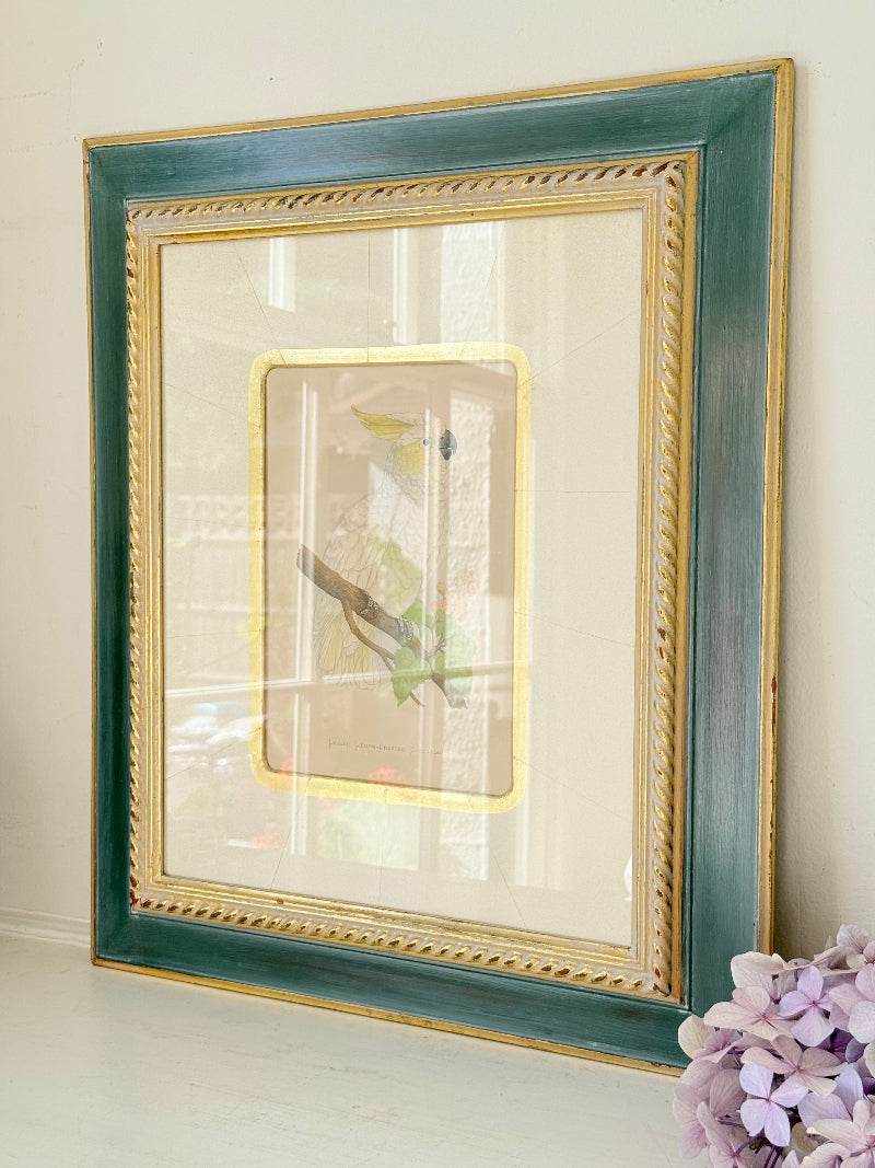 Framed cockatoo bird print in green and gold frame with mount, styled on white surface with vintage books