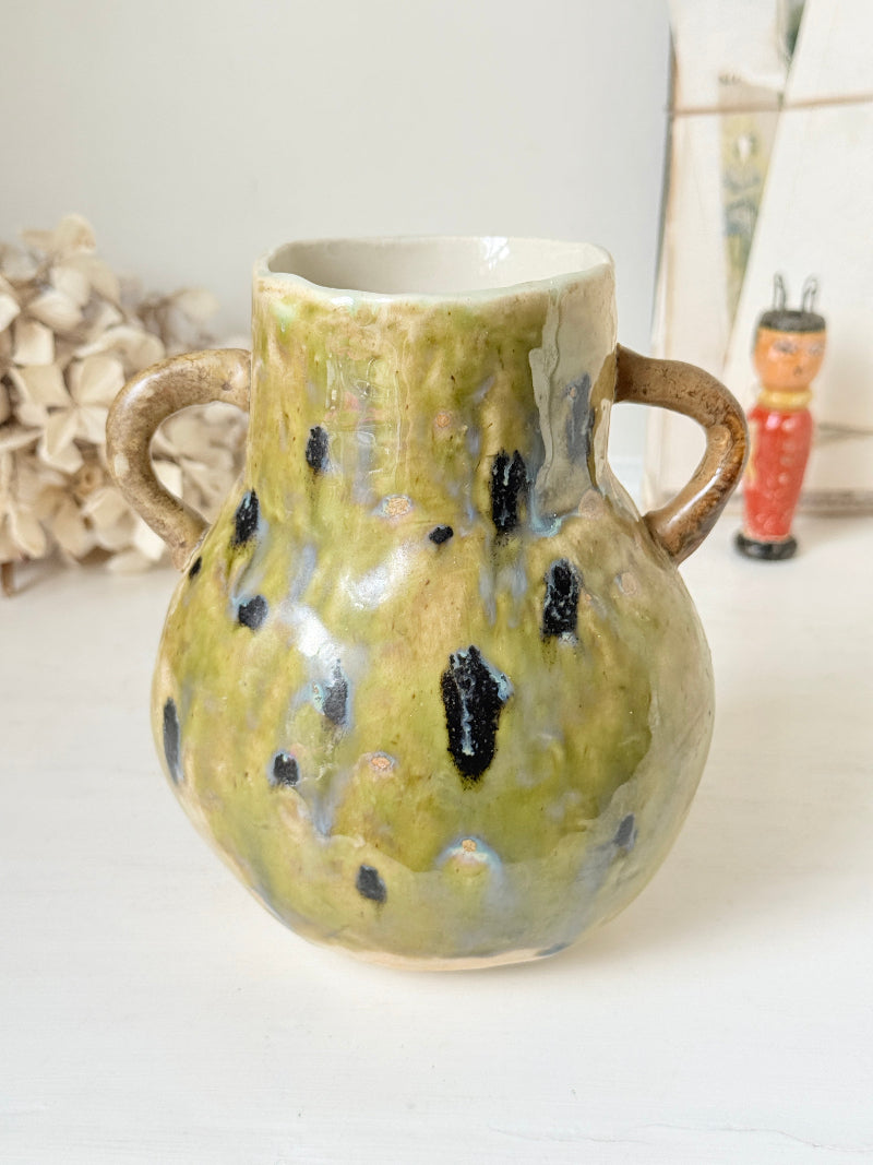 A handmade ceramic vase with two handles, featuring a textured glaze in olive green, beige, and black speckles, set against a softly blurred background with neutral decor.