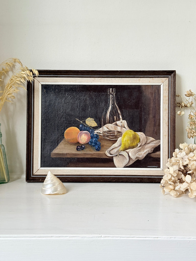 A framed still life oil painting on a white shelf, surrounded by dried decorative grasses and a small shell. The painting features an orange, grapes, a pear, and a glass bottle on a dark background.