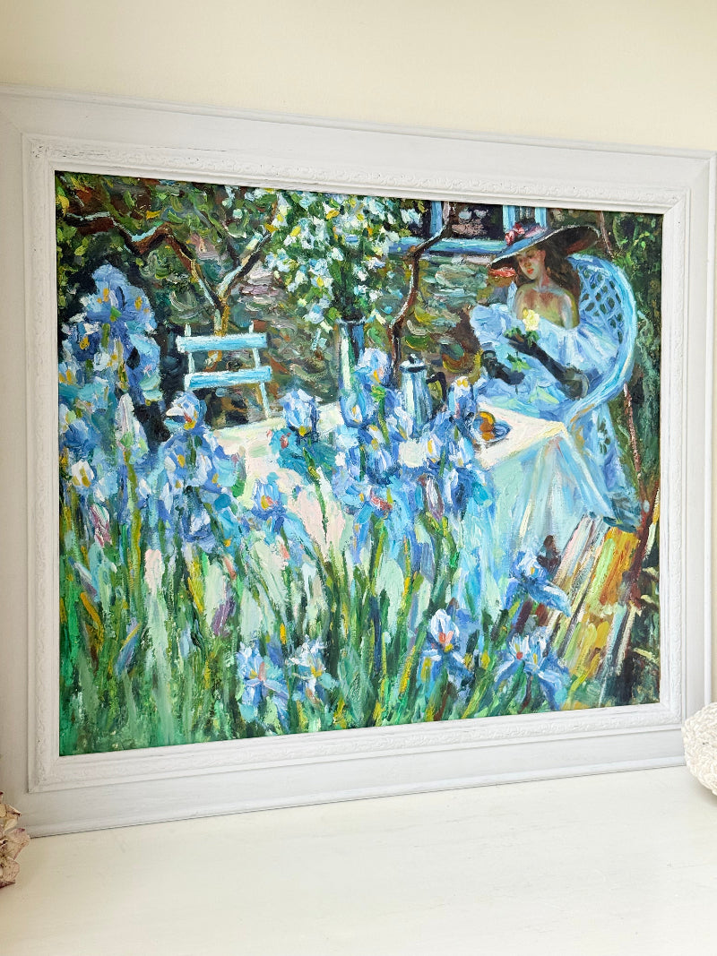 Framed impressionist-style oil painting of blue irises and garden scene, photographed on a white surface