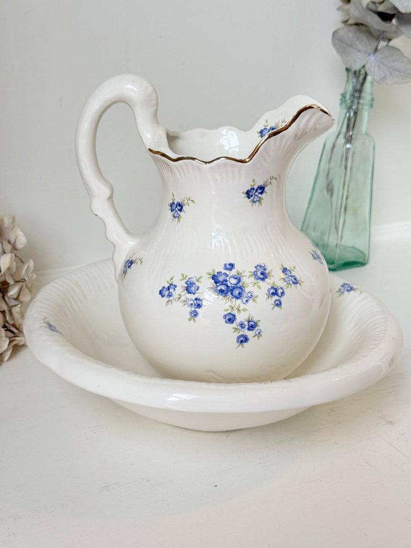 A white ceramic pitcher with blue floral patterns and a gold-trimmed fluted rim, shown on a matching white footed saucer. The pitcher features delicate blue rose clusters and is styled on a light surface with a green glass bottle and dried flowers in the background.