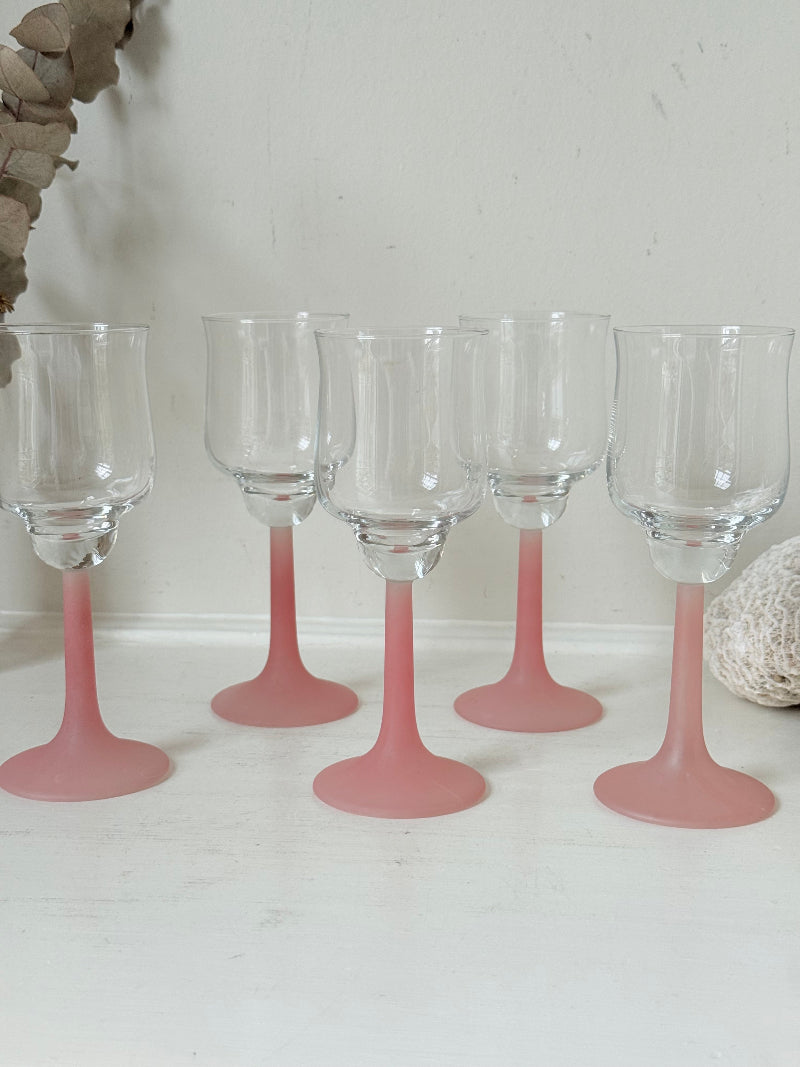 Set of five vintage wine glasses with clear bowls and pink stems photographed on a white surface.