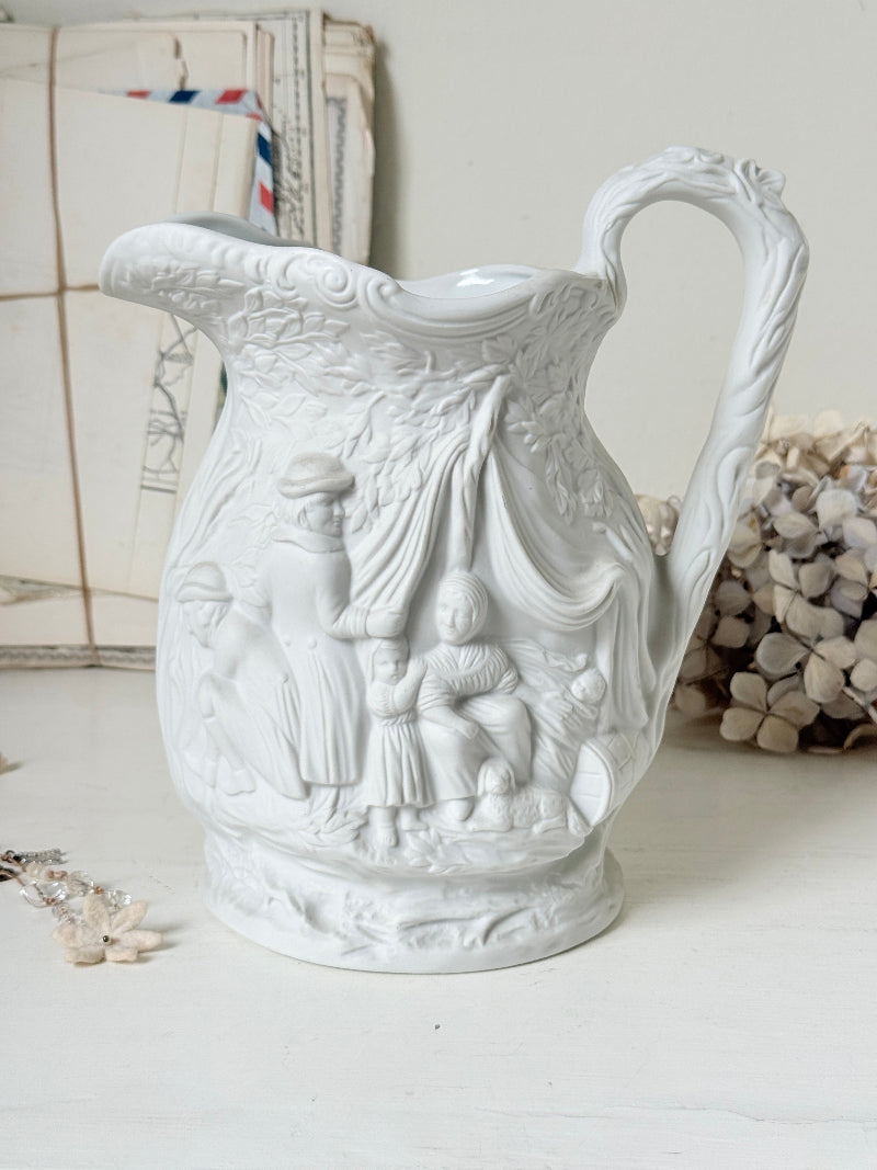 A white ceramic pitcher with intricate embossed classical figures and a draped scene, set against a softly styled background with vintage mail and dried flowers.