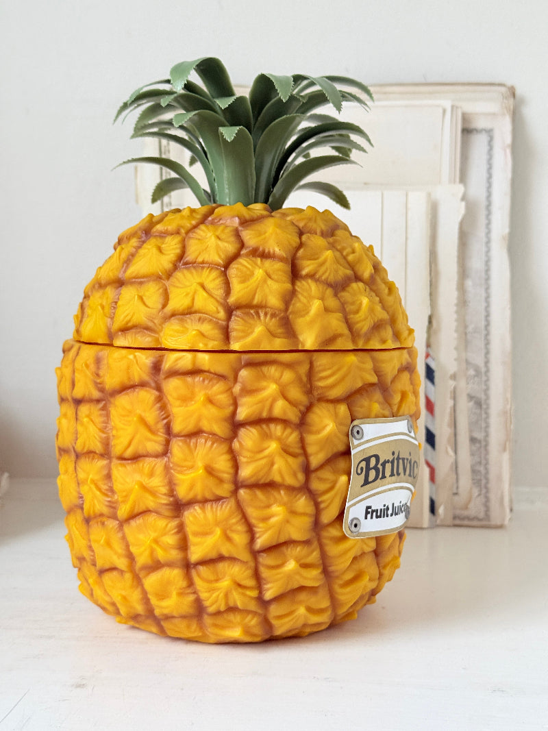 A novelty container shaped like a pineapple, with a textured yellow-orange body, green leafy top, and a small hanging label that reads &