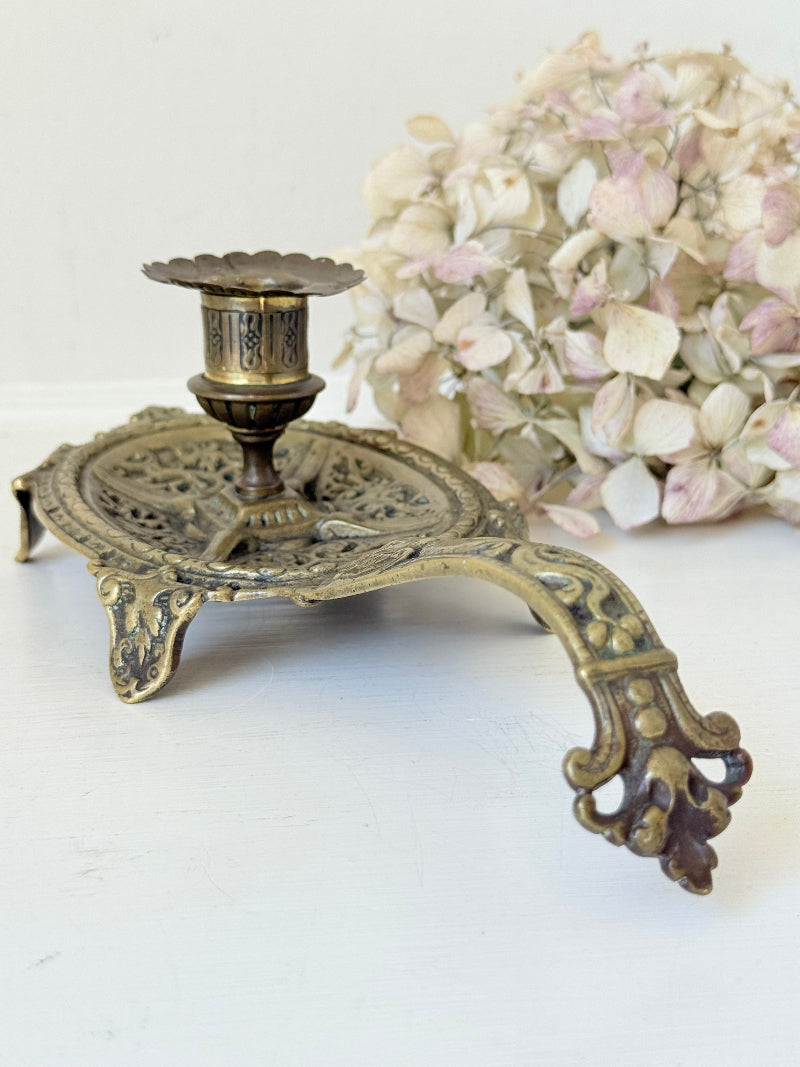Ornate vintage brass candlestick with decorative handle and footed base, photographed on a white surface