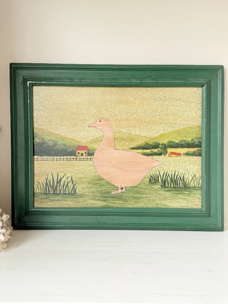 A framed folk art print featuring a pink goose standing in a green field with rolling hills and a small red house in the background, housed in a dark green wooden frame.
