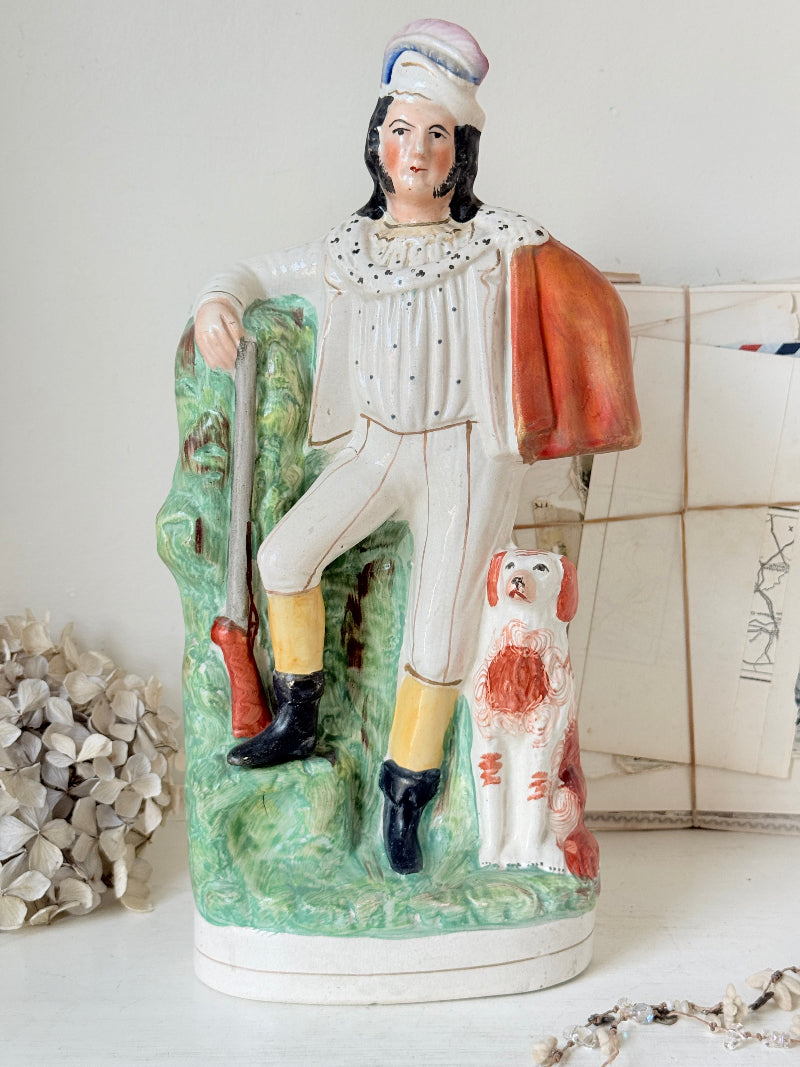 A vintage hand-painted ceramic figurine of a man in hunting attire leaning against a green tree, with a white dog at his side, displayed on a tabletop.