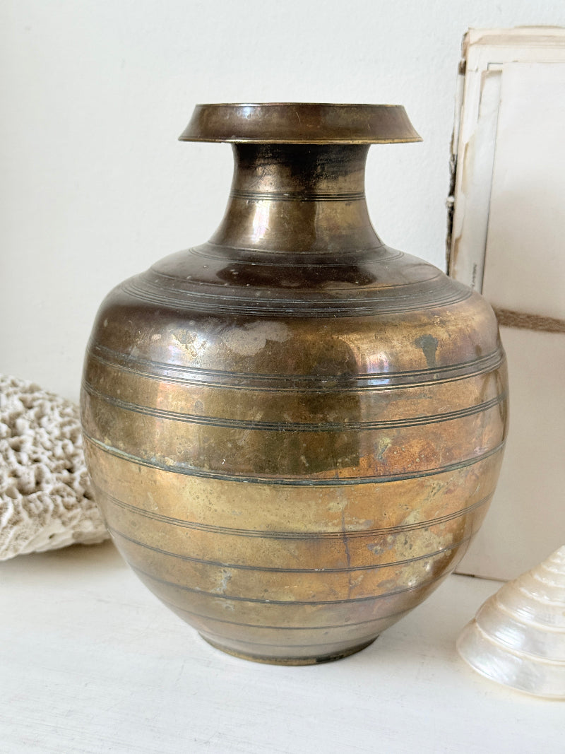 A vintage metallic bronze vase with horizontal ridges, displayed on a white surface next to decorative seashells.