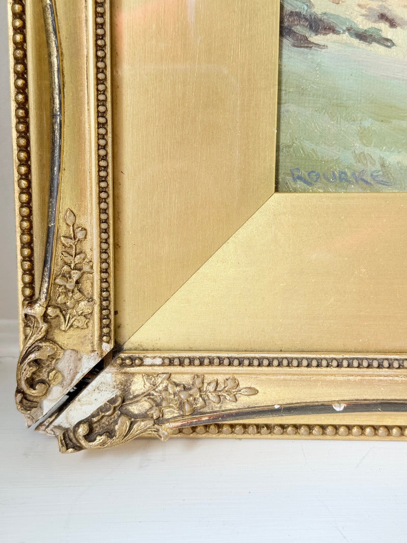 Framed oil painting of a rolling countryside landscape under a dramatic cloud-filled sky in an ornate gilt frame with wide mount.