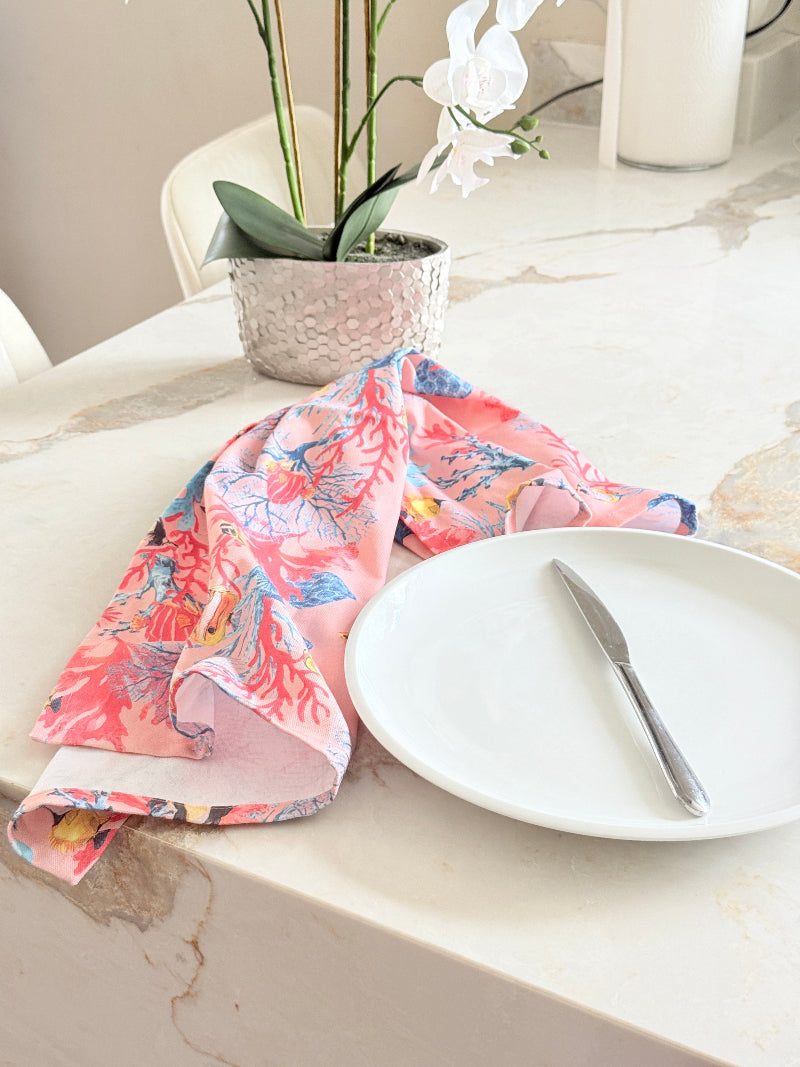 Warm peach coral and tropical fish print cotton tea towel with wooden forks on a marble surface beside cut glass