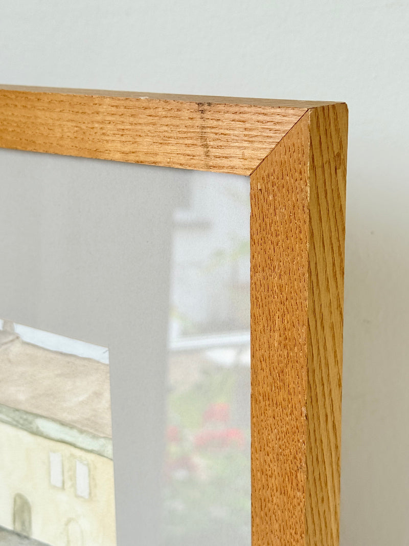 Framed neutral-toned village scene watercolour with figures in light oak-effect frame and pale grey mount, on white surface.